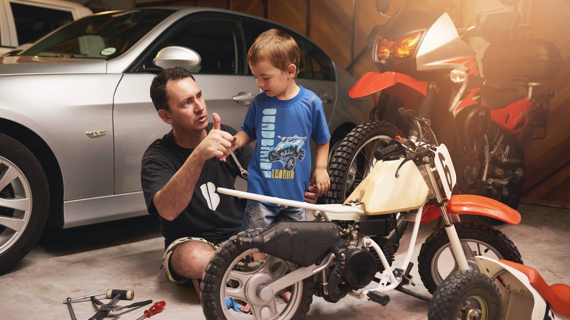 Padre enseñando a su hijo a arreglar su moto en el garaje de su casa en el cual tiene sus vehículos con un seguro combinado