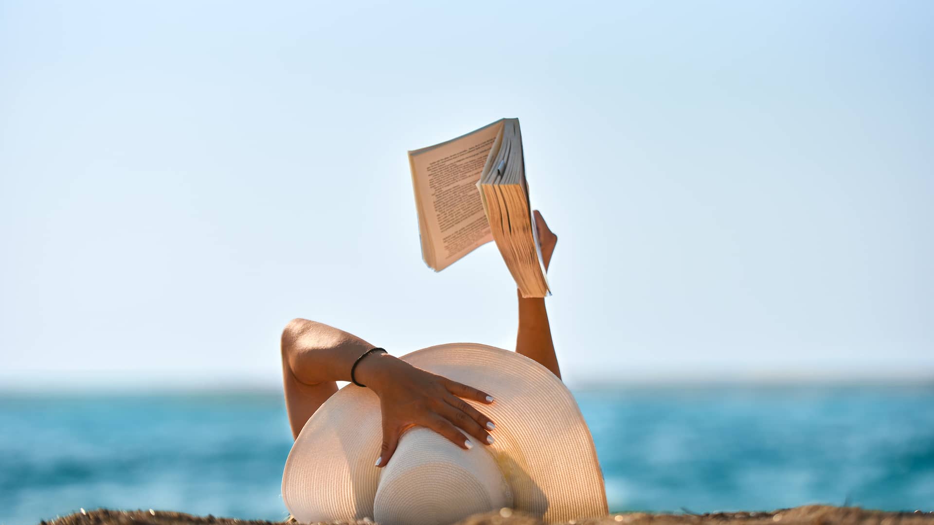 Mujer leyendo tumbada en la arena de la playa disfrutando de unas plácidas vacaciones con la contratación de su seguro de viaje